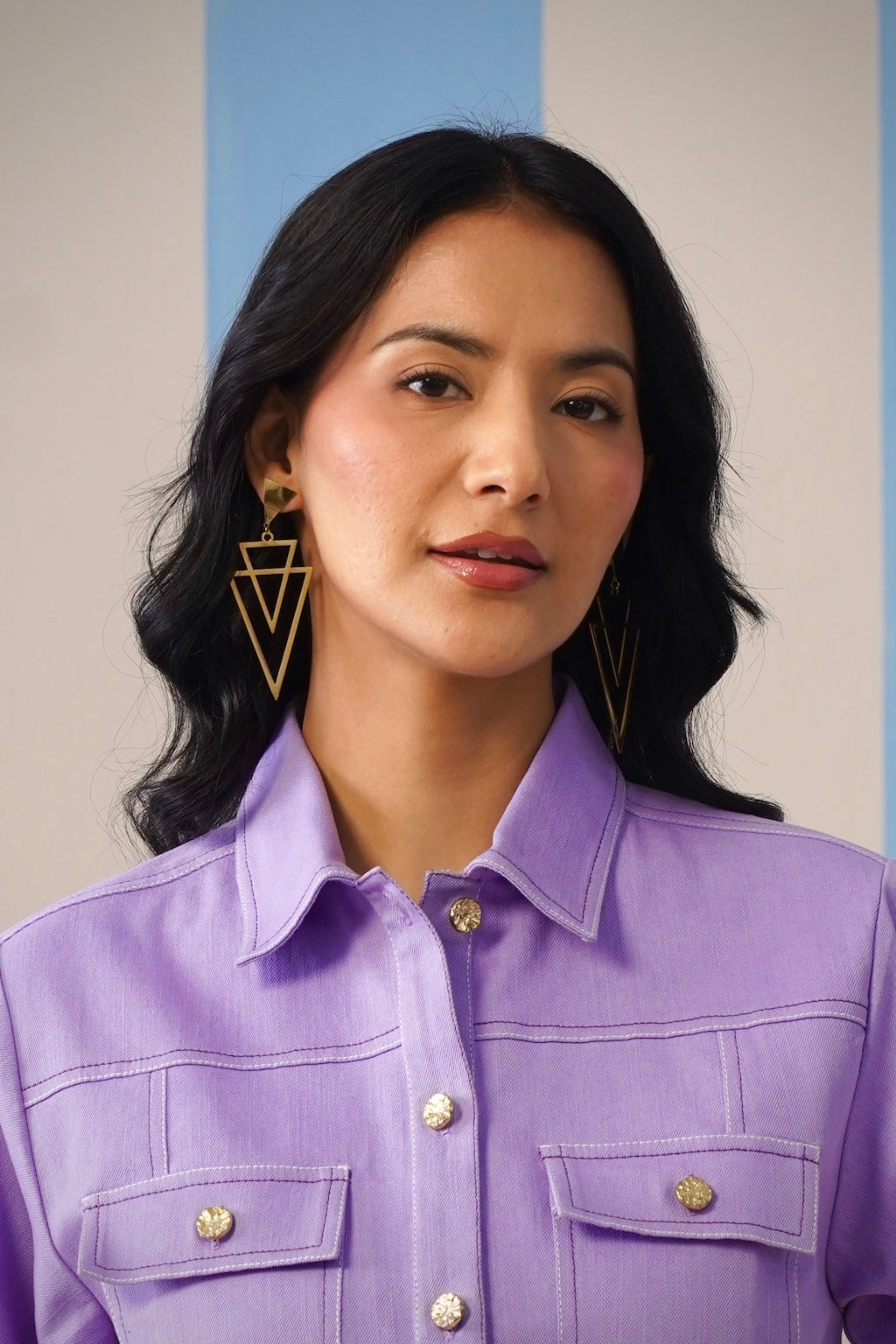 Woman wearing a purple shirt with gold buttons and earrings against a neutral background