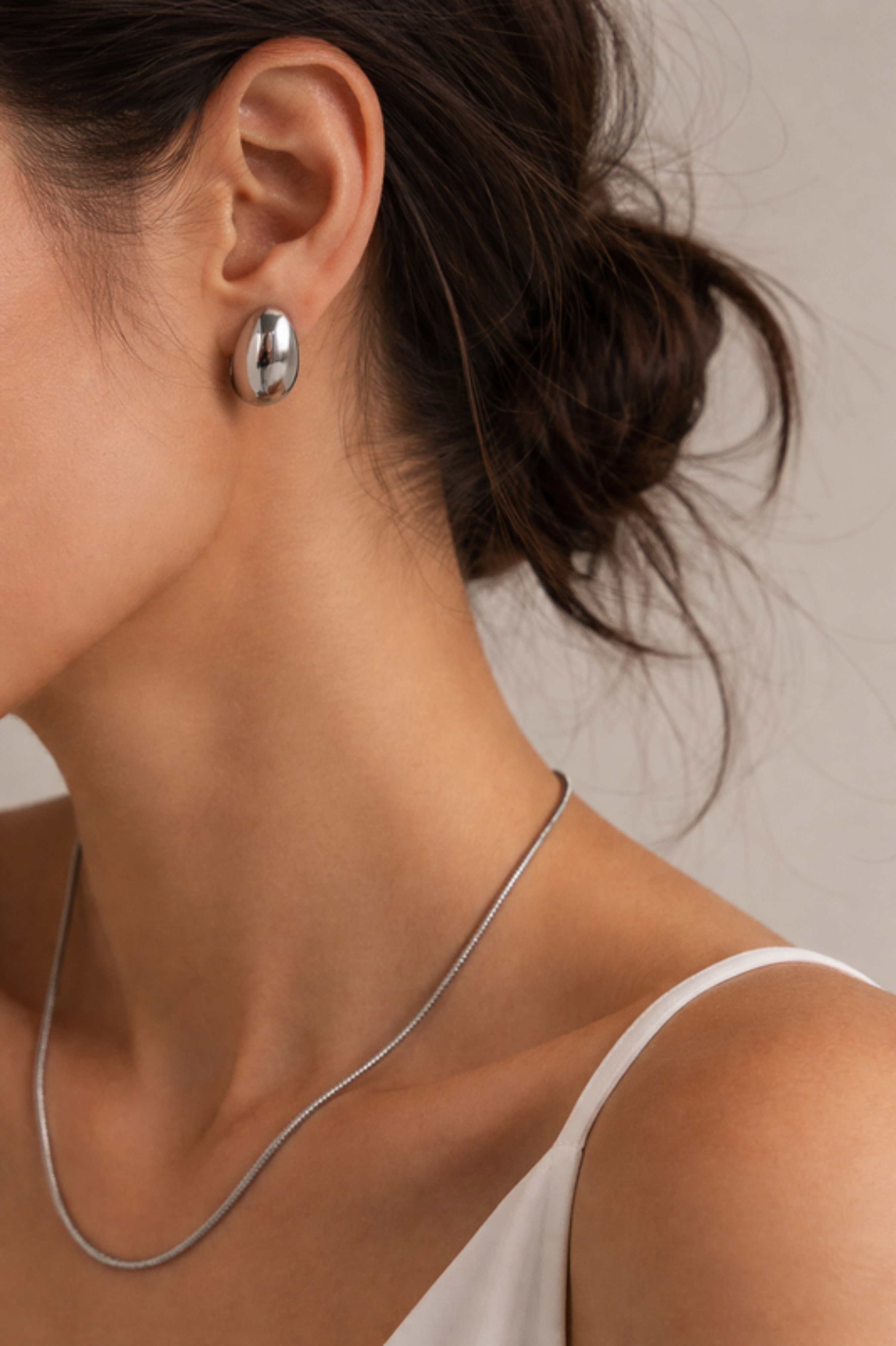 Close-up of a person wearing silver earrings and a necklace against a neutral background