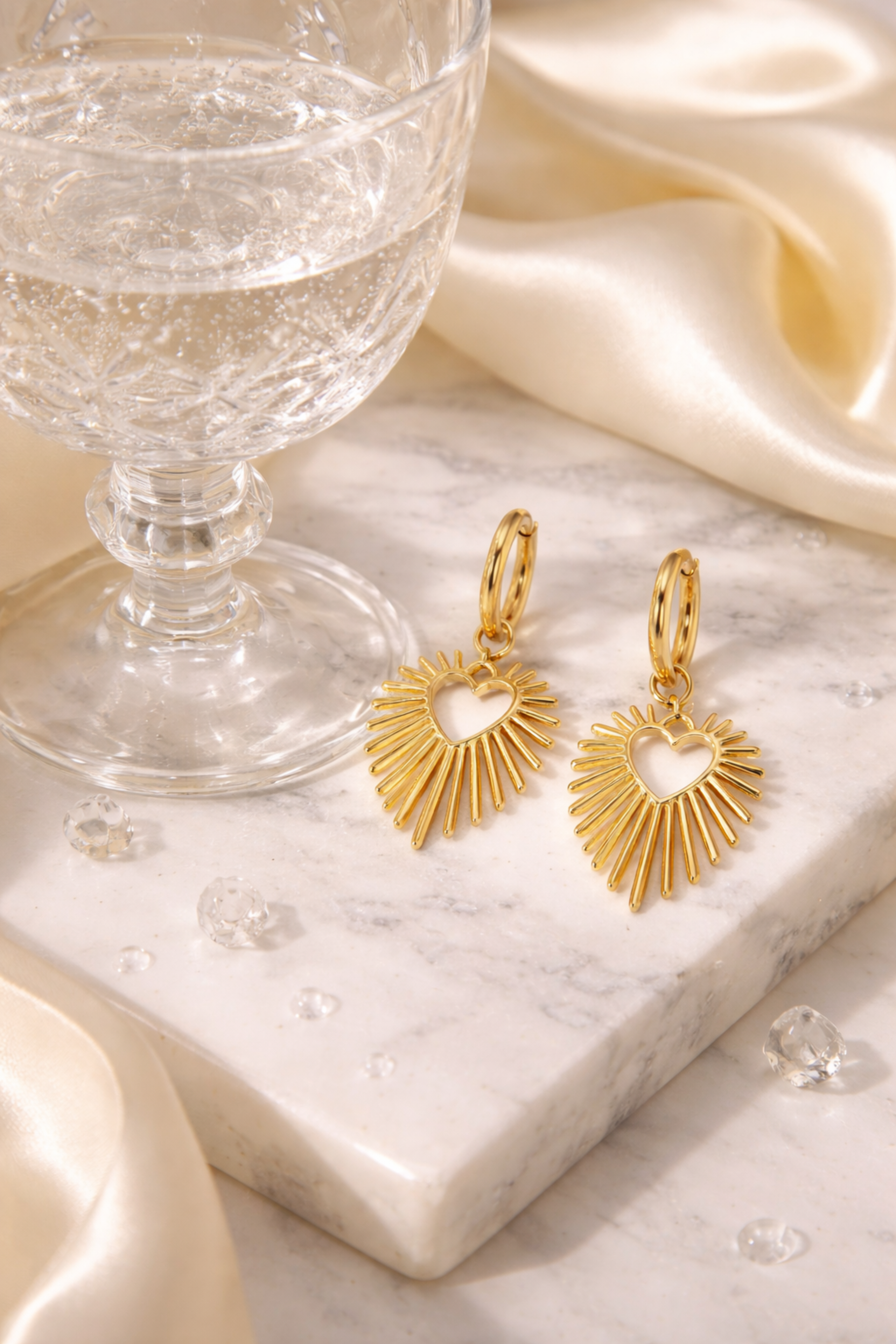 Gold heart-shaped earrings on a marble surface with a glass of water and fabric in the background.
