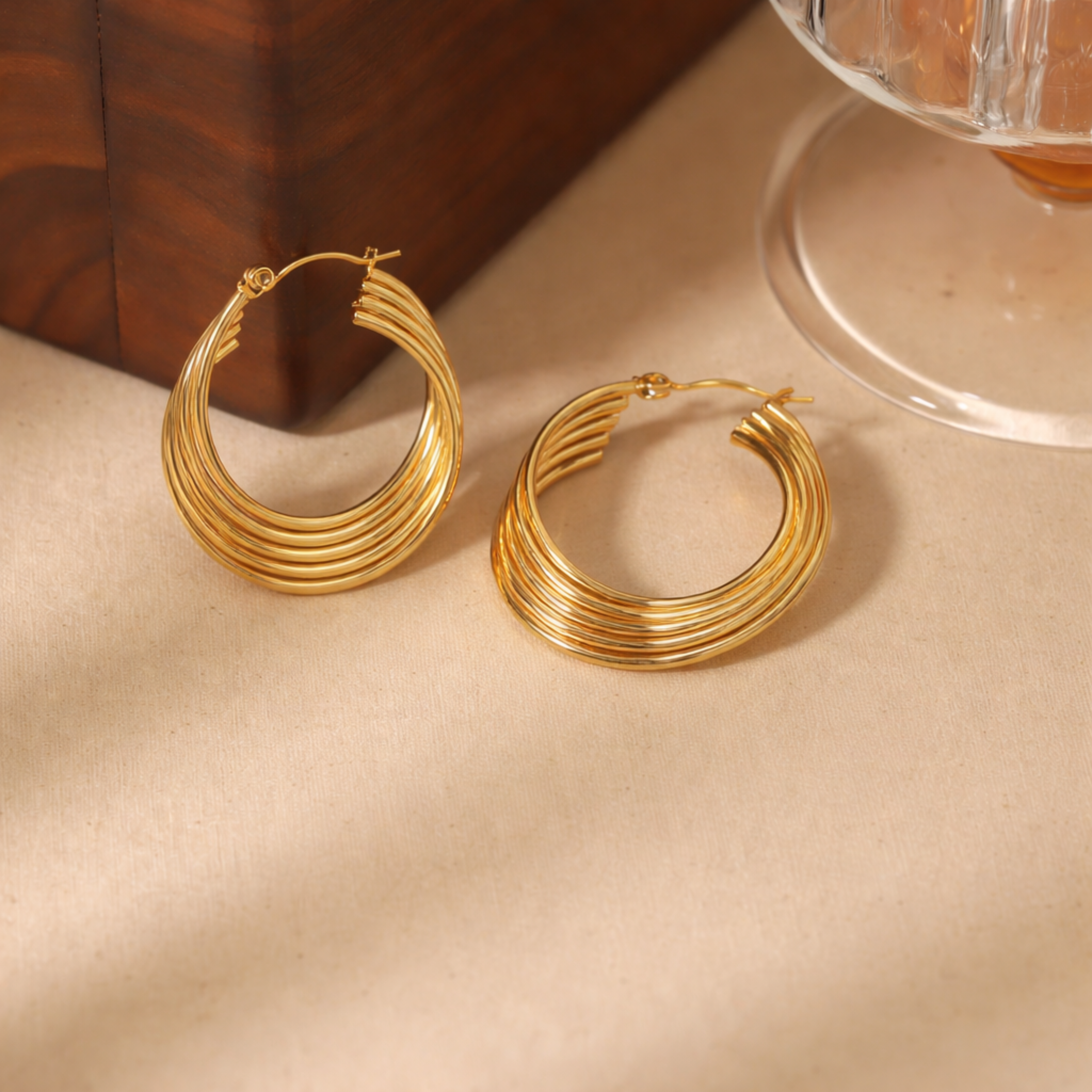 Gold hoop earrings on a beige surface with a wooden box and glass container in the background.