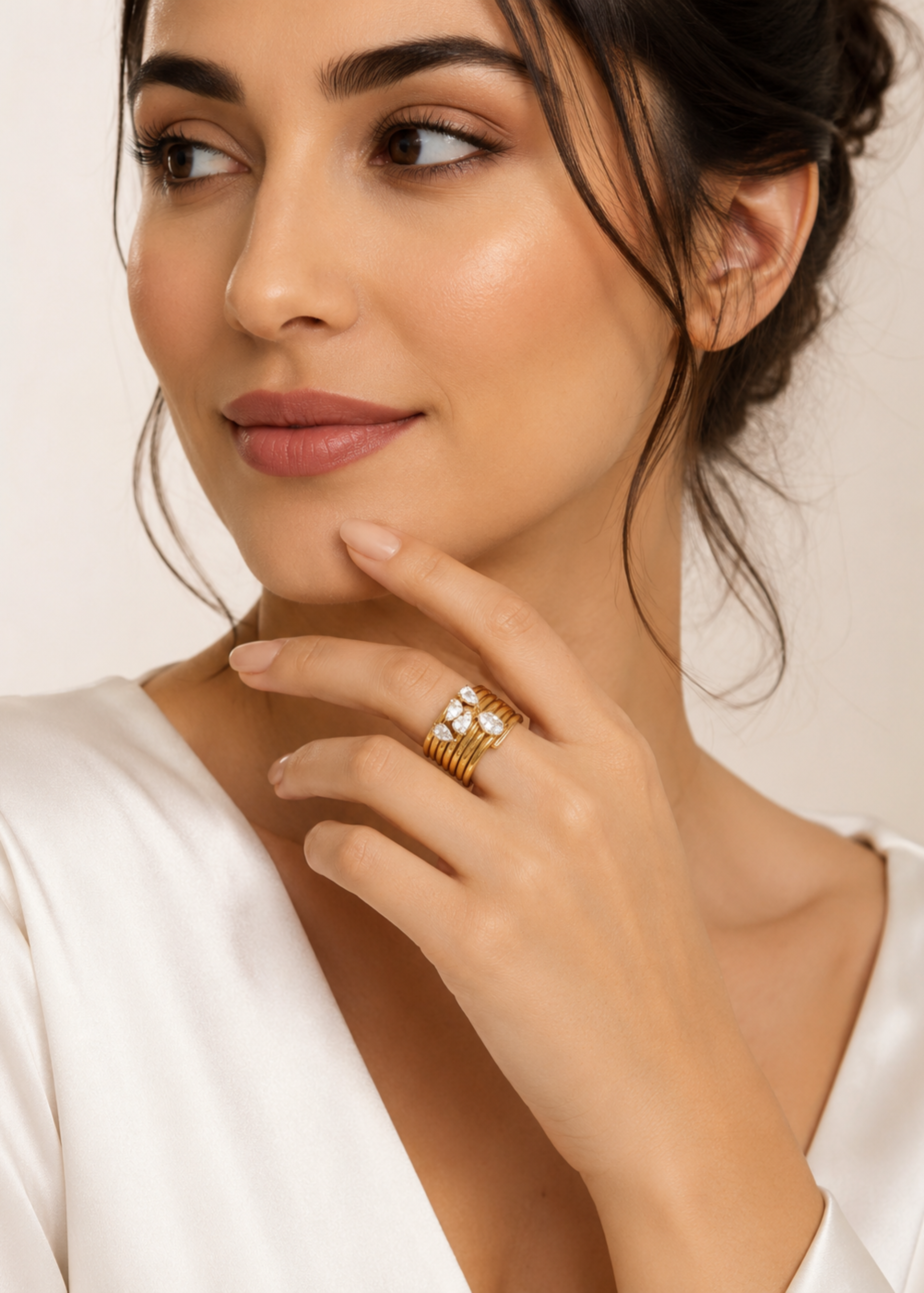 A close-up of a woman wearing a stack of gold rings, including one with a white stone cluster, against a soft beige background.