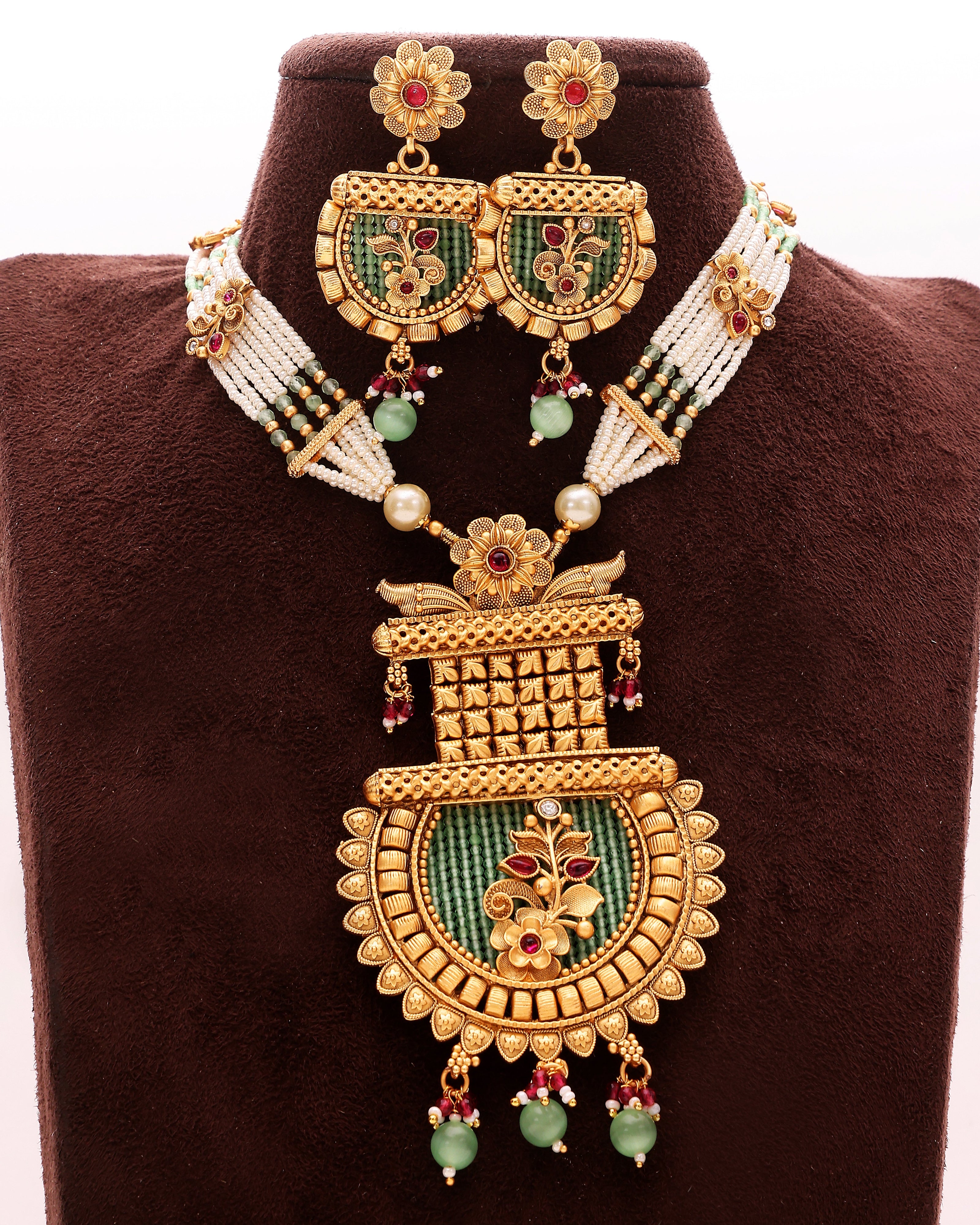 Ornate gold necklace with green stones and red gemstones on a brown velvet stand.