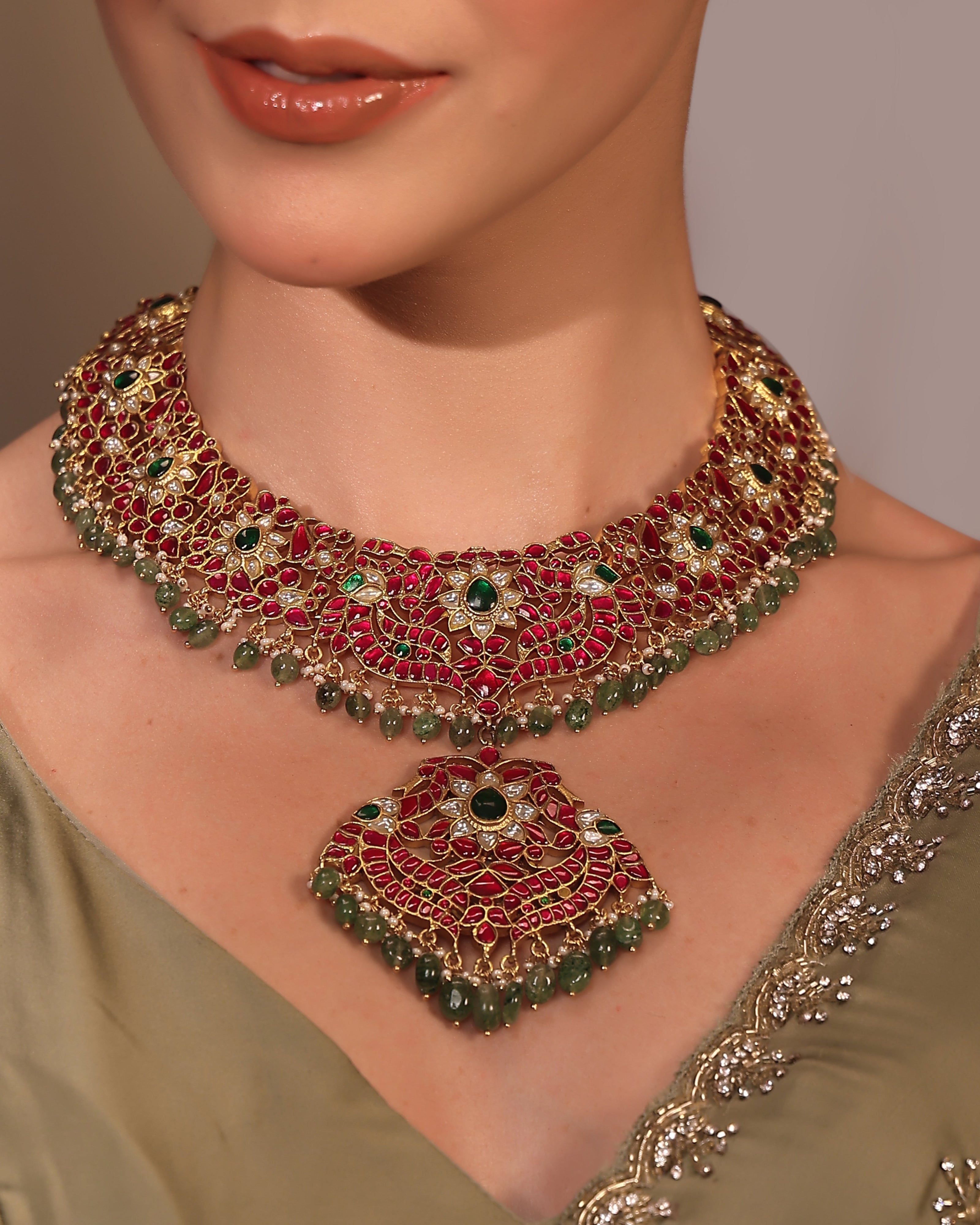 Close-up of a woman wearing an ornate necklace with red and green gemstones on a neutral background.