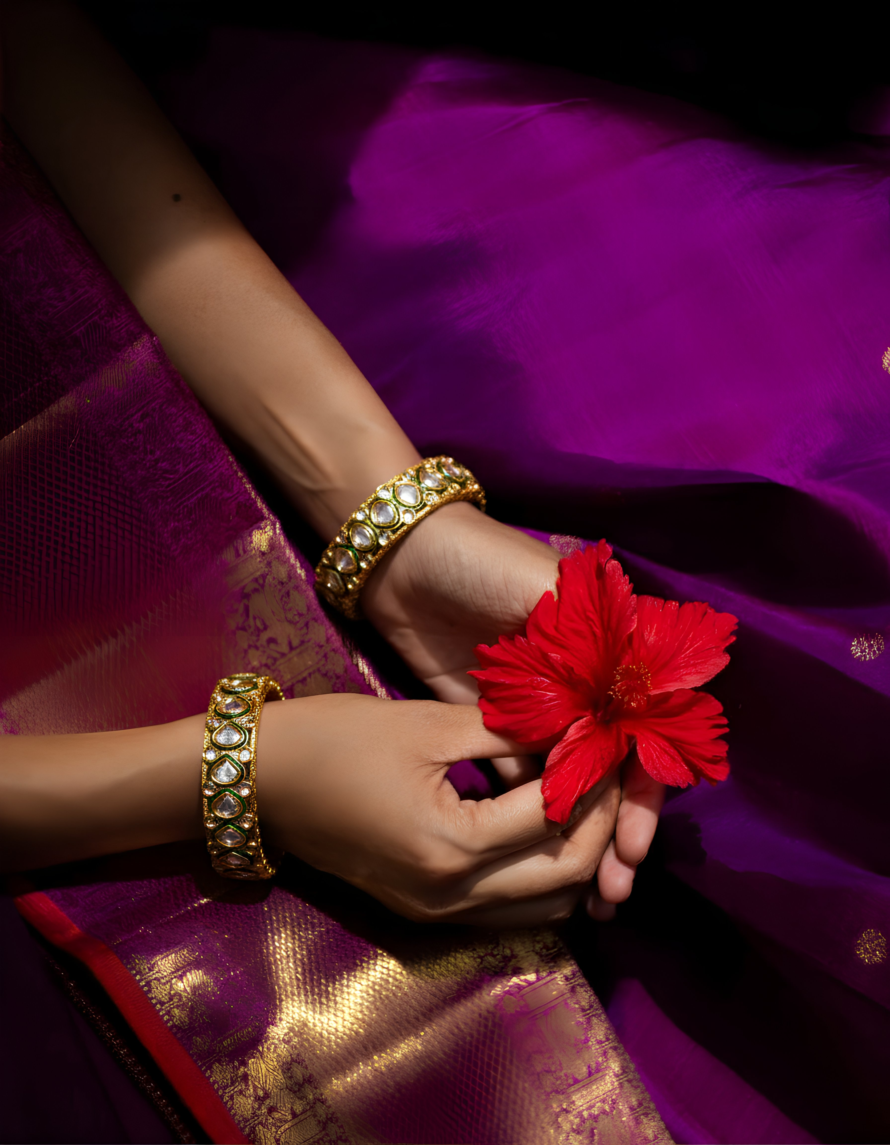 Person holding a red flower with gold bangles on a purple fabric background
