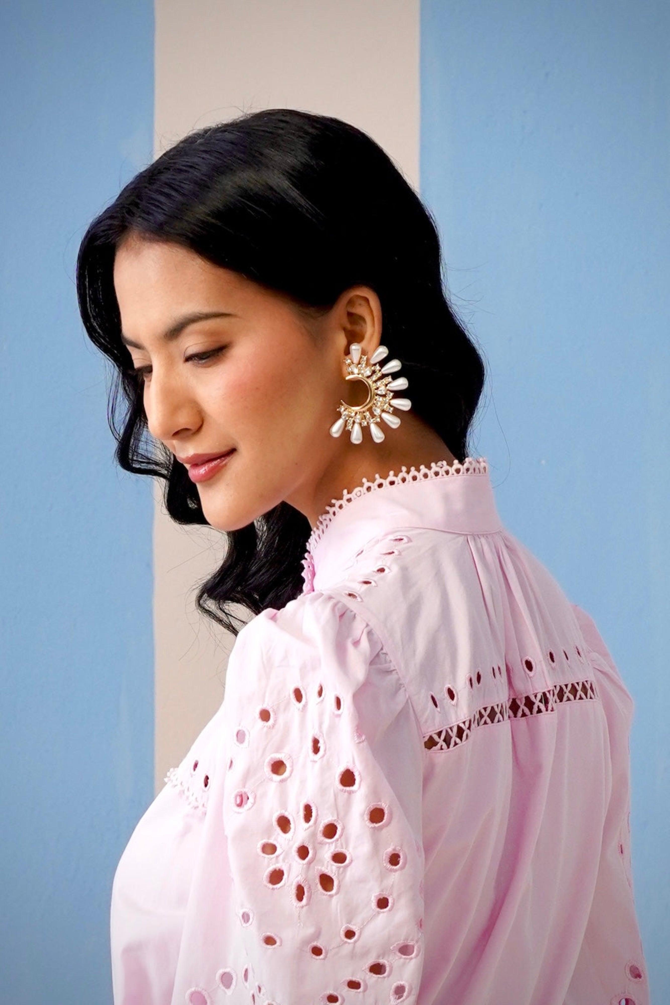 Woman wearing a white blouse with lace details and gold earrings against a light blue background