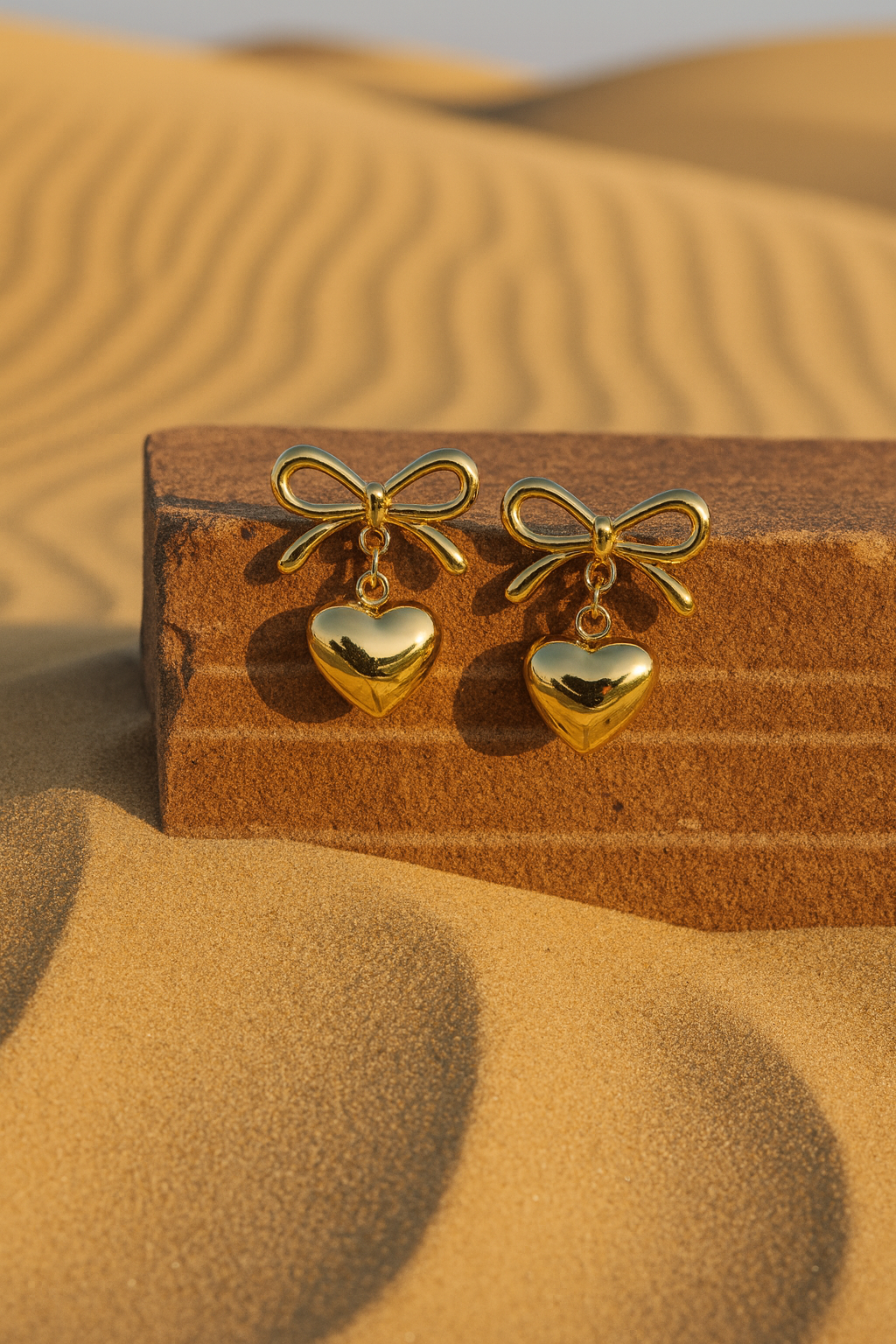Gold bow and heart earrings on a wooden block with sand dunes in the background