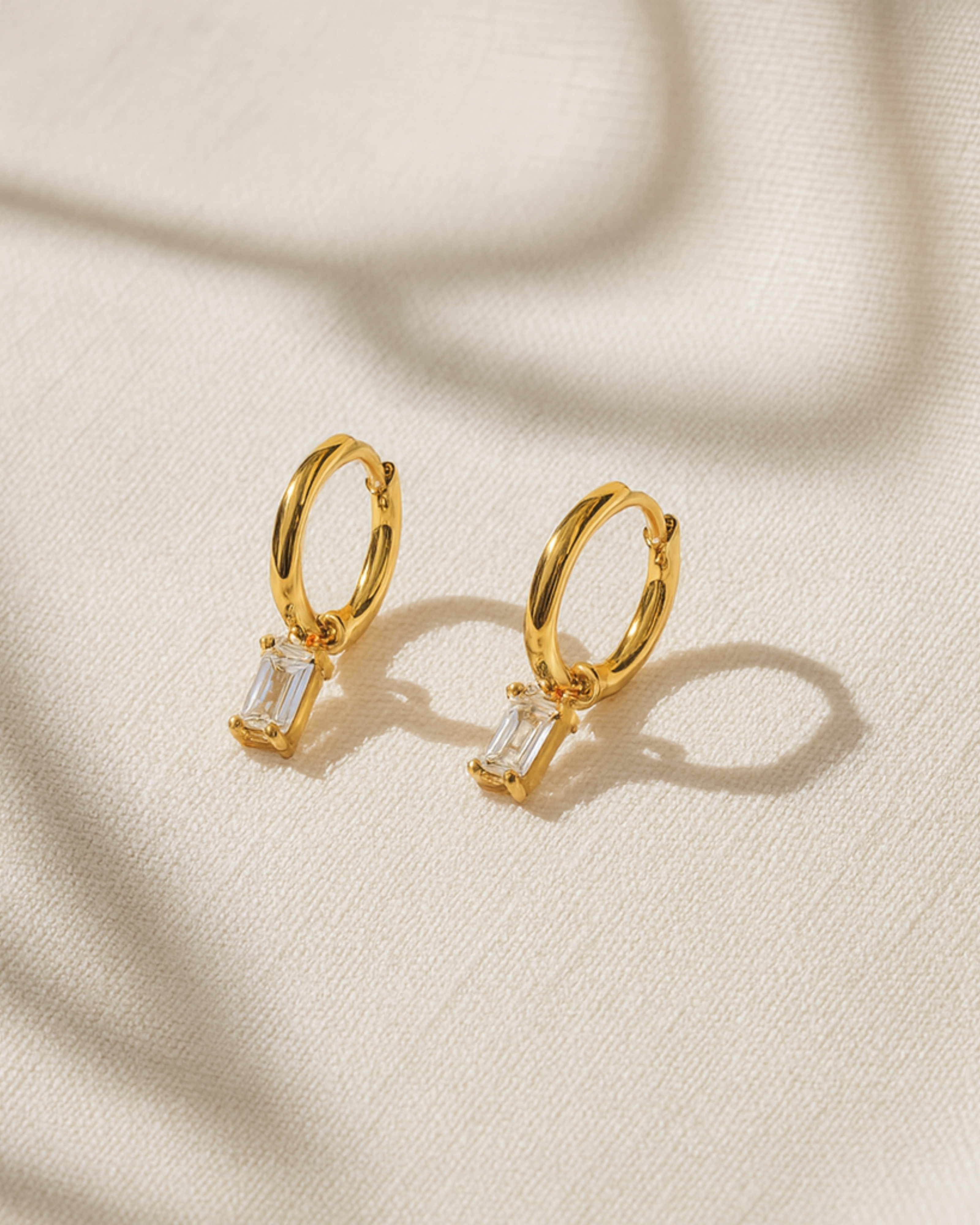 Gold hoop earrings with small rectangular gemstones on a beige fabric background