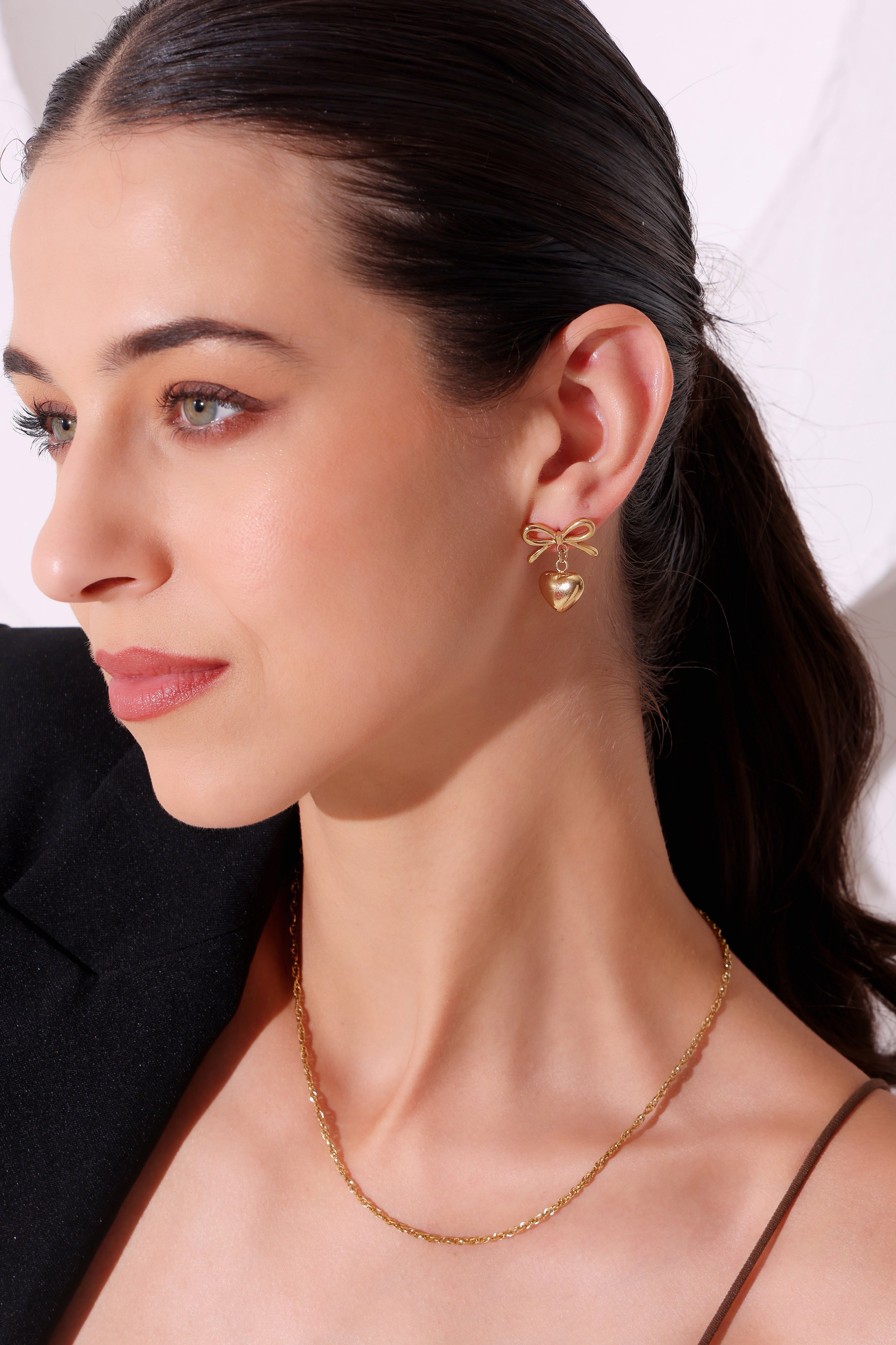 Woman wearing gold heart-shaped earrings and a delicate necklace against a white background
