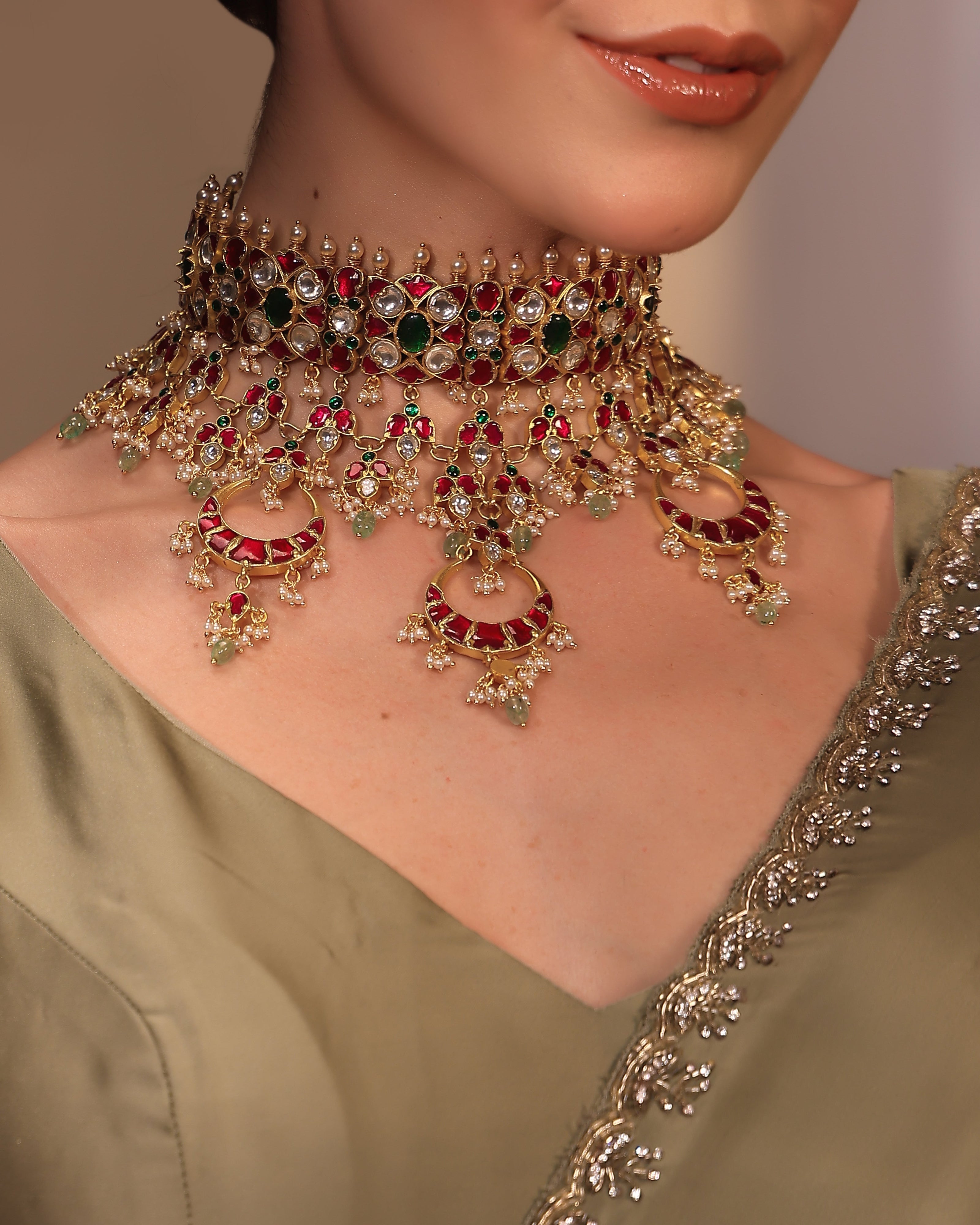 Close-up of a person wearing an ornate necklace with red and green gemstones on a plain background.