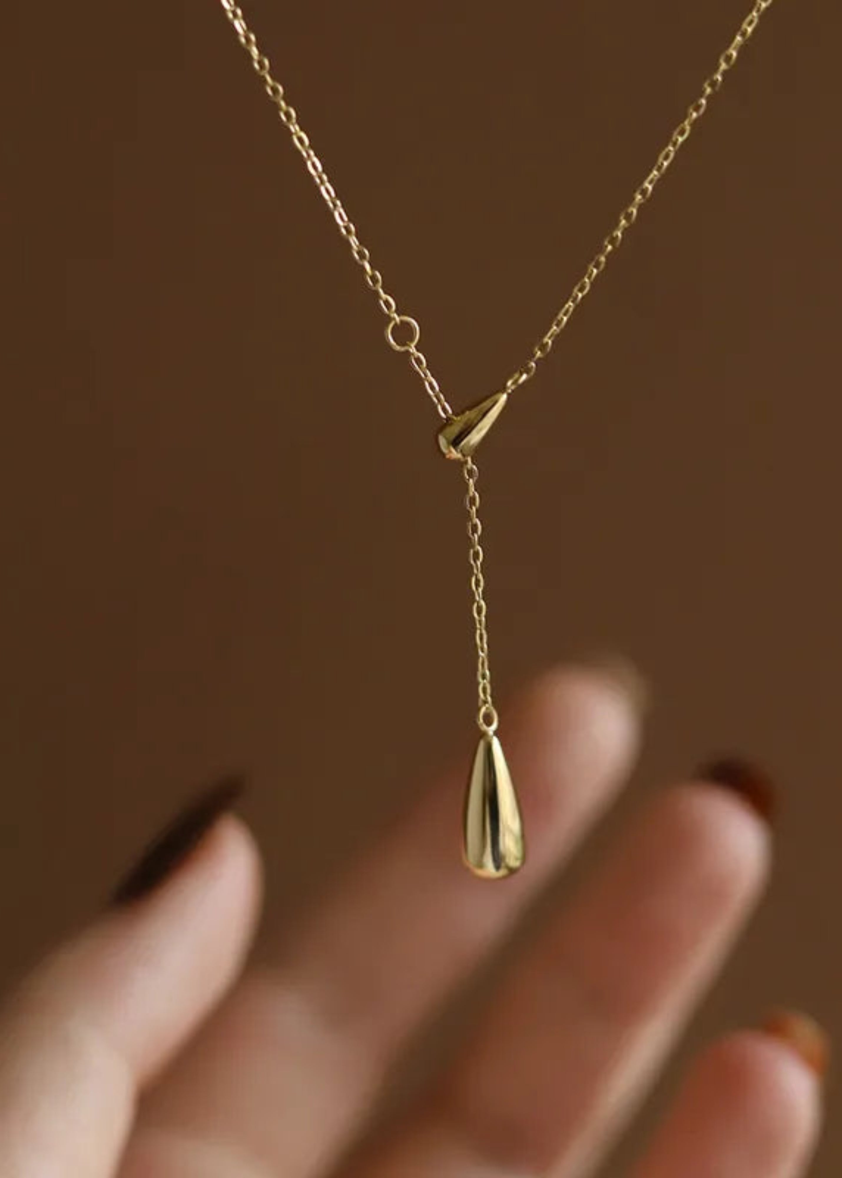 Gold necklace with a teardrop pendant held by a hand against a brown background